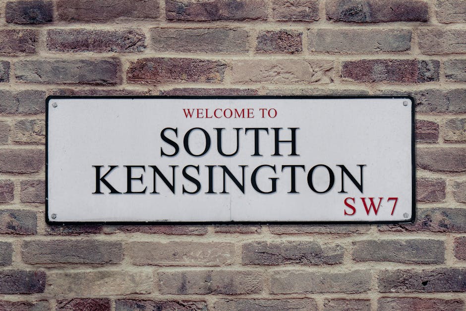 A rectangular white street sign mounted on a red brick wall displaying the address for South Kensington in the SW7 postal district, with the text 'WELCOME TO SOUTH KENSINGTON' printed in black and red. The sign is positioned at eye level, with visible mounting screws on each corner. The brick wall has a textured surface with varying shades of reddish-brown bricks, some slightly weathered. This scene could be part of a property exterior in a residential or commercial area in London, where house removals or relocation services such as those offered by Removal Van Kentish Town might operate, especially during home relocation or furniture transport processes.