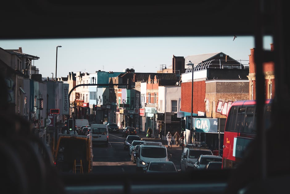 A view of a busy street in Kentish Town seen from inside a vehicle, with the window frame visible at the top and sides of the image. The street has several small shops with colorful signs and storefronts, including a barbershop and a retail store. Parked and moving cars fill the road, and pedestrians are walking along the pavement. In the foreground, the back of a large red and grey moving or delivery van managed by Removal Van Kentish Town is visible, parked close to the curb. The van’s rear doors are open, revealing empty interior space prepared for loading furnishings, boxes, or appliances in the home relocation process. The scene is lit by natural daylight, with clear visibility of the surrounding buildings, streetlamps, and the urban environment typical of residential and commercial mixed-use areas. This street scene illustrates a typical location for furniture transport and packing and moving activities involved in residential relocations in Kentish Town.