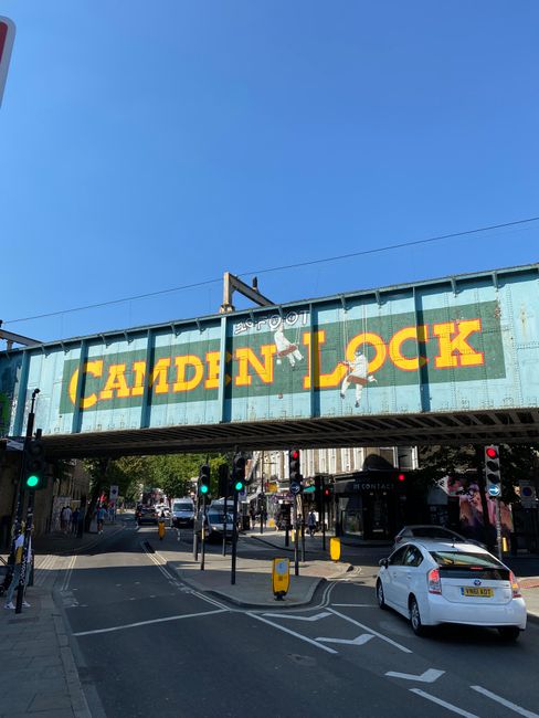 A large, weathered railway bridge painted in light blue with the words 'CAMDEN LOCK' in bright yellow letters spans over a busy street in Kentish Town. The bridge features a graffiti-style illustratio