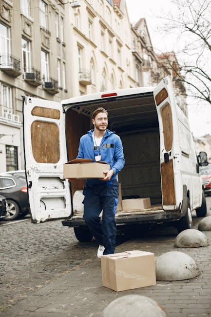 A professional mover from Removal Van Kentish Town is seen unloading cardboard boxes from a white cargo van parked on a residential street. The van's rear doors are open, revealing the interior used f