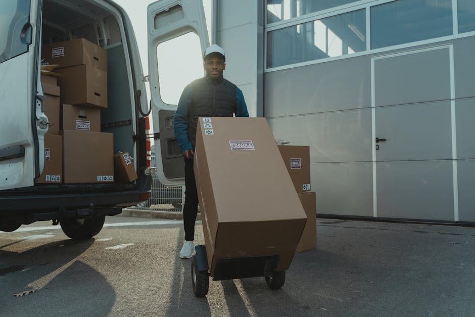 A professional mover from Removal Van Kentish Town is seen unloading cardboard boxes from a white cargo van parked on a residential street. The van's rear doors are open, revealing the interior used f