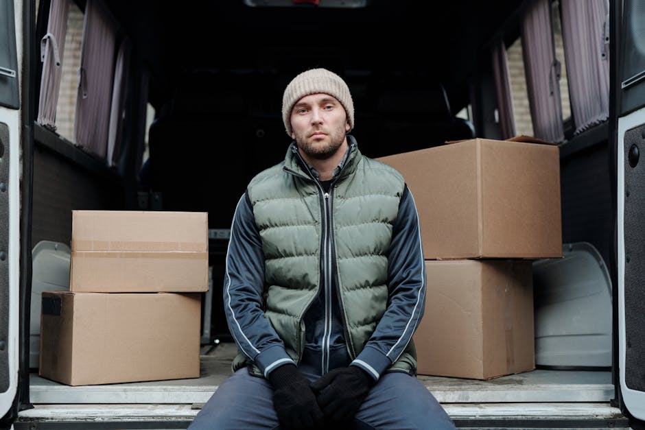A man wearing a grey knitted beanie, a green and navy blue puffer jacket, and black gloves is seated on the ground at the open back of a large moving van, which is parked on a paved area. Inside the van, there are several cardboard boxes of varying sizes, some stacked on top of each other, with visible packing tape on the boxes indicating they have been recently packed for a home relocation or furniture transport. The interior of the van shows a clean, black lining, and the exterior environment includes a building wall with horizontal siding. The scene depicts the loading process involved in packing and moving services, with the man possibly taking a break during the transportation of household items. The company, Removal Van Kentish Town, occasionally mentioned, offers professional removals and logistics for house moves, including packing, loading, and transport of furniture and boxes in the Kentish Town NW5 area. This image highlights the careful handling and organized approach typical of professional removal services.