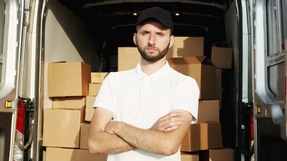 A young man with a serious expression, wearing a white polo shirt and a black cap, stands inside the rear of a moving van loaded with stacked cardboard boxes of various sizes, some wrapped in plastic. The boxes occupy most of the space behind him, indicating the packing process involved in home relocation or furniture transport. The van's interior is illuminated, showing a glimpse of the vehicle’s dark ceiling and side walls. The man is positioned in front of the boxes with his arms crossed, suggesting a moment during the loading process for a house removals service. The image is taken outdoors on a paved surface, with part of the van's open door visible on the right, providing context for a house or apartment move. This scene visualizes the logistical aspects of packing, loading, and transport involved in professional removals services, as provided by Removal Van Kentish Town, supporting the services listed under the 'removals' category on the Fortess Road in Kentish Town NW5.