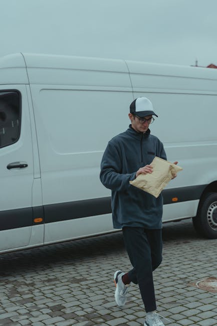 A man wearing a grey knitted beanie, a green and navy blue puffer jacket, and black gloves is seated on the ground at the open back of a large moving van, which is parked on a paved area. Inside the van, there are several cardboard boxes of varying sizes, some stacked on top of each other, with visible packing tape on the boxes indicating they have been recently packed for a home relocation or furniture transport. The interior of the van shows a clean, black lining, and the exterior environment includes a building wall with horizontal siding. The scene depicts the loading process involved in packing and moving services, with the man possibly taking a break during the transportation of household items. The company, Removal Van Kentish Town, occasionally mentioned, offers professional removals and logistics for house moves, including packing, loading, and transport of furniture and boxes in the Kentish Town NW5 area. This image highlights the careful handling and organized approach typical of professional removal services.