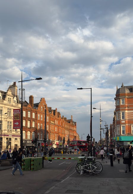 A view of a busy street in Kentish Town with a mix of historic brick buildings lining the roadside under a partly cloudy sky. In the foreground, a bicycle is parked next to a green plastic barrier near the pavement. Pedestrians are walking along the sidewalk, some carrying bags, as a red double-decker bus moves through the intersection. Several street lamps and traffic signs are visible, along with a few parked cars. The scene captures the urban environment typical of house removals and furniture transport in the area, highlighting the busy street scene where Removal Van Kentish Town might facilitate home relocation or moving services, with vehicles and loading processes likely taking place nearby.