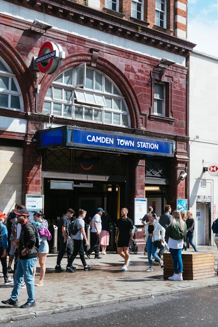 A group of pedestrians, including individuals carrying backpacks and shopping bags, standing outside the entrance of Camden Town Station, a red brick building with a blue sign displaying 'Camden Town Station'. The station features arched windows, a round underground sign, and a partially open entrance door. The scene is set on a paved sidewalk with some people waiting or preparing to enter the station, alongside a nearby wooden bench on the right, with clear daylight illuminating the area. The image captures a typical busy scene during daytime at a popular London underground station, relevant to house removals or relocation services such as those provided by Removal Van Kentish Town, which includes planning for parking permits and adhering to Camden Council rules.