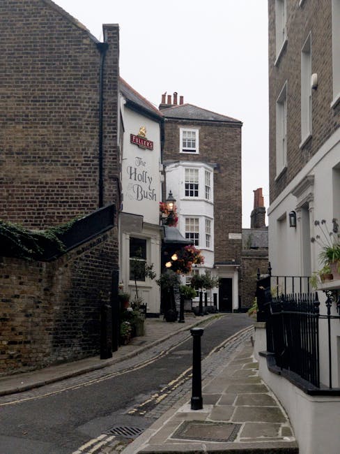 A narrow residential street in Kentish Town featuring a steep, cobbled pavement lined with brick and stucco buildings. On the left side, a white pub called 'The Holly Bush' displays hanging flower baskets and potted plants outside its entrance. On the right, a white building with black railings and potted greenery is visible. The street has black bollards along the edge of the pavement to prevent vehicle overrun. In the distance, a removal van is partially visible, parked along the street, with members of Removal Van Kentish Town engaged in loading or unloading furniture and boxes wrapped in cardboard, plastic, and fabric. The scene is captured during daytime with natural light illuminating the area, emphasizing the urban environment suitable for home relocation and furniture transport processes based on typical moving services.