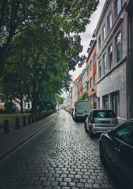 A wide cobbled street lined with historic stone-built terraced houses featuring pitched roofs and multiple chimneys. The houses have white-framed windows and small front gardens bordered by low stone walls. A black street lamp is visible on the left side near a sidewalk, and a person can be seen walking in the distance. The scene is illuminated by soft daylight with a partly cloudy sky overhead. The setting reflects a typical residential area suitable for house removals and furniture transport, and the image may encompass scenes relevant to the logistics of home relocation on Kentish Town Road, including access via narrow streets. Removal Van Kentish Town operates in such environments, assisting with packing, loading, and transporting belongings through these historic streets.
