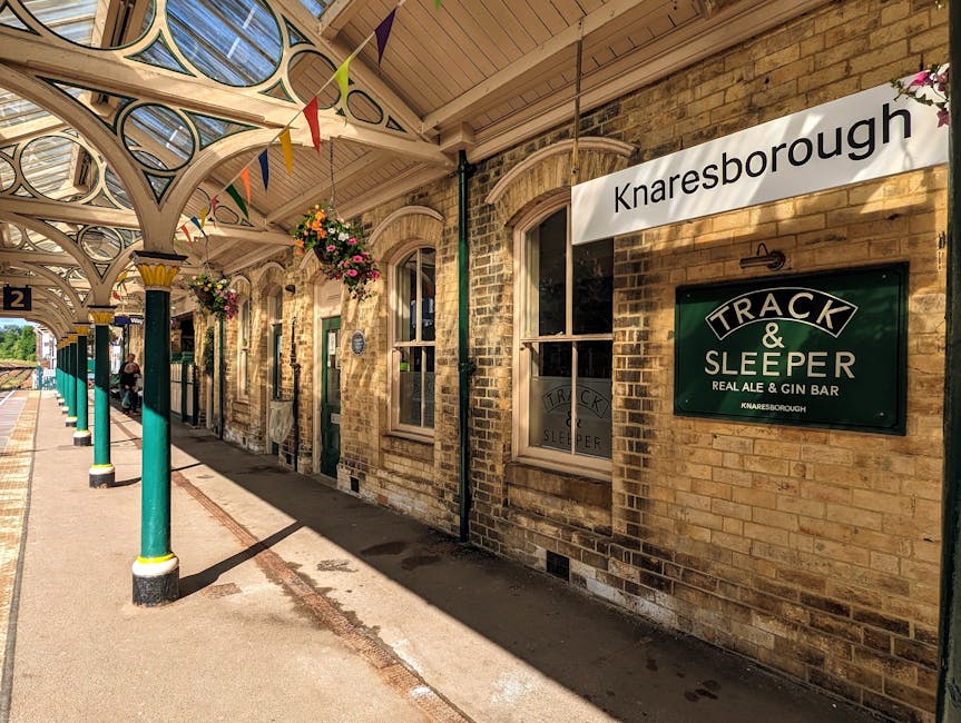 Photograph of Knaresborough train station platform during daytime, showing a row of brick station buildings with arched windows and decorative trim. The platform has green and yellow painted support columns with white decorative arches overhead, and hanging flower baskets with colorful flowers are suspended from the roof. A sign with the station name 'Knaresborough' is mounted on the brick wall next to a green board advertising 'Track & Sleeper Real Ale & Gin Bar.' The platform edge features a yellow tactile paving strip for safety, and a train is visible at the far end of the platform. The scene is well-lit with natural sunlight, highlighting the textures of the brickwork and the station's outer environment. This setting relates to house removals and short-distance moves, illustrating the transportation and logistics involved in furniture transport and packing during home relocation, supported by Removal Van Kentish Town.
