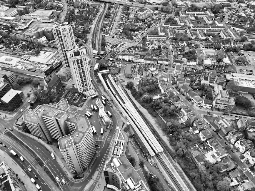 A black and white aerial photograph of an urban area showing tall office buildings, smaller residential houses, and roads. The image captures a section of a city with a railway line running through the middle, alongside a station platform where several trains are parked, indicating a busy transport hub. Surrounding the station, there are high-rise and mid-rise buildings with varying architectural styles. Streets filled with vehicles and buses are visible, along with pavements and smaller streets branching off. In the lower part of the image, a curved road with cars and trucks is seen, while the background shows a dense pattern of houses, trees, and additional roads. This view exemplifies a typical city environment suitable for home relocation and furniture transport services. As part of the services offered by Removal Van Kentish Town, this aerial view underscores the importance of precise logistics and effective packing and moving processes involved in house removals within the city.