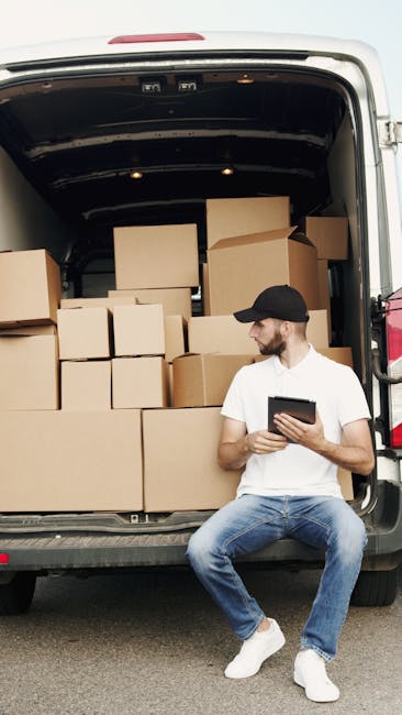 A man wearing a white t-shirt, black baseball cap, and blue jeans is seated on the edge of an open van's loading area, which is filled with stacked cardboard boxes of various sizes, some partially covered with protective plastic wrap. The boxes are arranged on the pavement, with some resting against the interior wall of the van, indicating a home relocation or furniture transport process. The man is holding a tablet device, possibly coordinating the packing or loading activities. Inside the van, the dark interior is visible, with additional boxes and packing materials. The scene captures the loading process typical of professional removals, such as those provided by Removal Van Kentish Town, during scheduled moving services involving packing, transportation, and logistics management for residential moves.