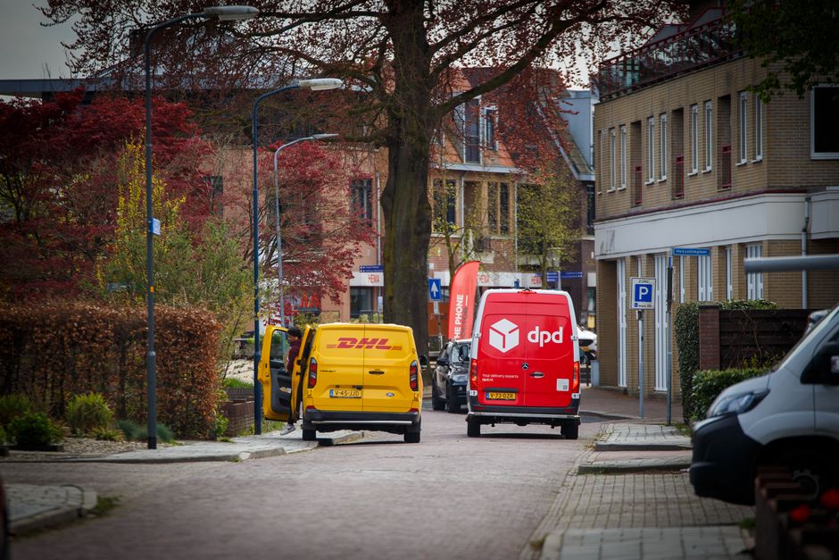 The image shows a residential street scene during daytime with two delivery vans from DHL and DPD parked on the pavement in front of a brick building. The yellow DHL van has its rear door open, with a person partially visible inside or near the vehicle, possibly involved in loading or unloading items. The red DPD van is parked directly behind the DHL van, with no visible activity around it. The street is lined with parked cars on both sides and features a tree with red and green foliage, indicating an autumn setting. To the right of the image, a portion of a white car is visible, and a parking sign indicating designated parking zones is mounted on a pole near the building. Street lamps and small landscaping bushes enhance the scene’s residential environment. The setting suggests a home relocation or furniture transport operation being carried out by Removal Van Kentish Town, with the vans positioned for efficient loading or unloading, aligned with professional moving and packing procedures in an urban neighbourhood.