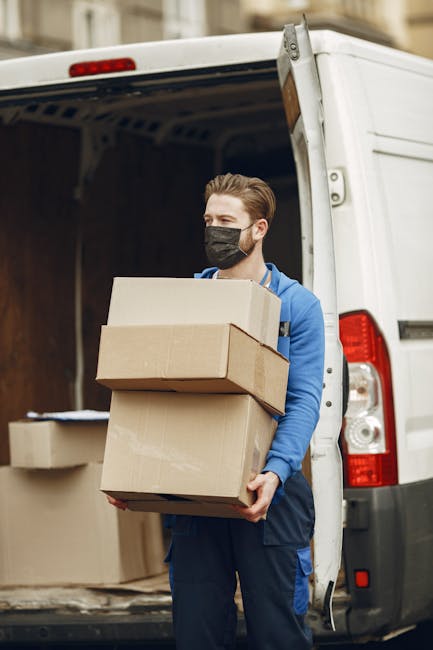 A man wearing a white t-shirt, black baseball cap, and blue jeans is seated on the edge of an open van's loading area, which is filled with stacked cardboard boxes of various sizes, some partially covered with protective plastic wrap. The boxes are arranged on the pavement, with some resting against the interior wall of the van, indicating a home relocation or furniture transport process. The man is holding a tablet device, possibly coordinating the packing or loading activities. Inside the van, the dark interior is visible, with additional boxes and packing materials. The scene captures the loading process typical of professional removals, such as those provided by Removal Van Kentish Town, during scheduled moving services involving packing, transportation, and logistics management for residential moves.