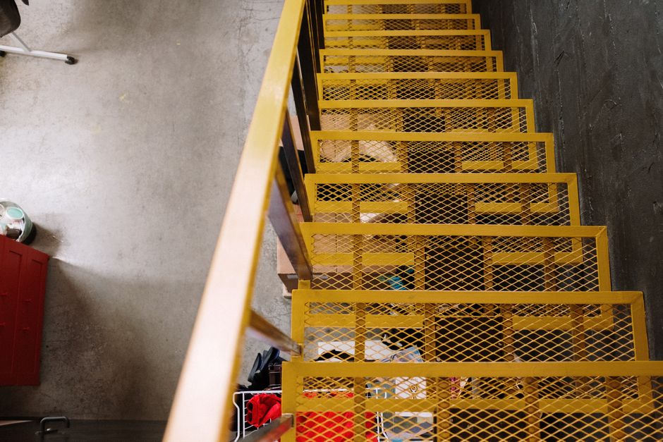 A high-angle view of a narrow staircase with yellow metal mesh steps and black side panels, situated inside a building during a home relocation. The staircase is used for moving furniture and boxes, with several cardboard boxes and wrapped items visible on the steps and at the bottom, indicating packing and loading activities by Removal Van Kentish Town. To the left of the staircase, a section of a concrete floor and a red storage unit are visible, along with part of a small white object on the floor. The environment is lit with natural and artificial light, highlighting the industrial look of the staircase, which is positioned adjacent to a dark-walled interior. The staircase facilitates furniture transport and packing during a stair access process, supporting efficient house removals, with a focus on handling items through limited spaces.
