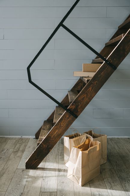 A close-up view of a staircase inside a property, showing dark wooden steps with a light wood finish on the edges, and a black metal handrail forming a geometric pattern against a light grey paneled wall. At the base of the stairs, there are several brown paper shopping bags filled with empty or partially filled items, placed on a wooden floor. The scene captures the process of home relocation, with the stairs ready for furniture transport and packing materials positioned nearby, illustrating a typical step in the removals process carried out by Removal Van Kentish Town. The lighting is natural and soft, highlighting the textures of the materials involved in the packing and moving activities.