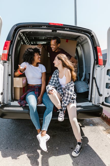 Two young women sit inside the open back of a loading van parked on a street in daylight, smiling and engaging in conversation during a home relocation process. The woman on the left has curly black hair, wears a light blue T-shirt, and has a red wristband, while the woman on the right has long, light brown hair, a white sleeveless top, and a plaid shirt tied around her waist. Behind them, the van's interior contains cardboard boxes, some sealed with packing tape, and moving blankets partially visible. The moving van, associated with Removal Van Kentish Town, is positioned with its rear door fully open, revealing the loading area designed for the transport of household furniture and belongings. This scene captures the moment of packing and moving, emphasizing the collaborative effort involved in furniture transport and home relocation, while highlighting the professional handling of packing materials like cardboard boxes and protective covers, and the logistics of loading items onto the van in preparation for a house removal.