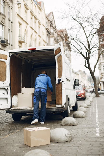 A person wearing a blue jacket and dark trousers is loading large white cardboard boxes into the back of a white removal van parked on a paved city street. The van's rear doors are wide open, revealing an interior lined with wooden panels. Several additional cardboard boxes, some taped and others unwrapped, are placed on the pavement nearby. The street is lined with multi-storey Victorian-style buildings with white facades and ornate balconies, and leafless trees are visible along the sidewalk. The scene is illuminated by natural daylight, with a few parked cars visible in the background. This image illustrates a home relocation process, involving furniture and box loading for a professional removals service, such as provided by Removal Van Kentish Town, supporting efficient moving logistics.