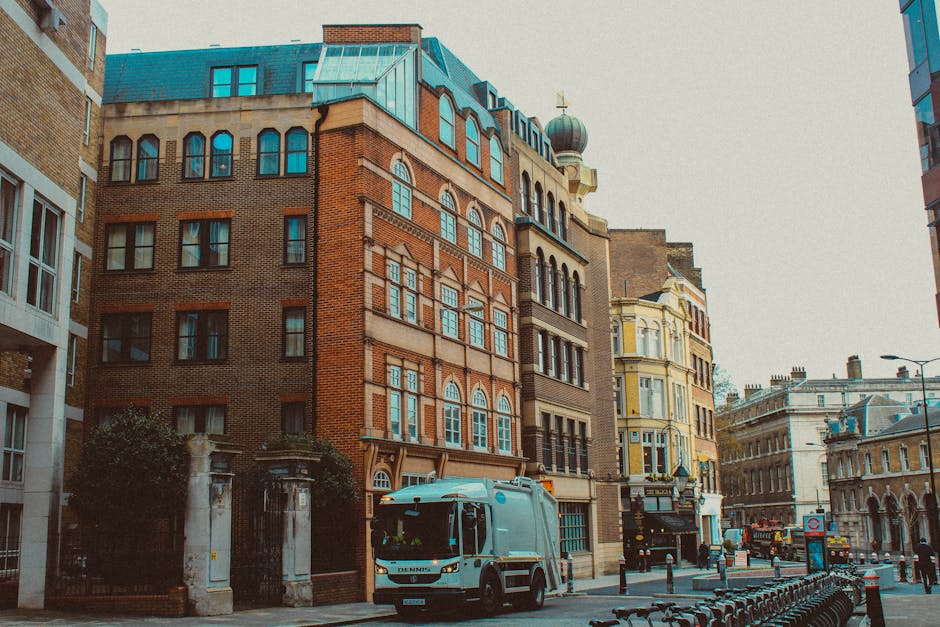 A street scene in Kentish Town featuring a collection of multi-storey buildings with diverse architectural styles, including red brick facades and decorative window designs. A blue Removal Van Kentish Town is parked on the pavement near a building entrance, with its rear doors open, suggesting active loading or unloading. Several cardboard boxes, some wrapped in plastic or fabric, are positioned on the pavement close to the vehicle, indicating packed household or office items ready for a home relocation. The loading process involves furniture and boxes being handled for transport into or out of the van. Nearby, equipment such as trolleys and straps are visible, supporting safe and efficient movement of items. The environment is well-lit, with ambient daylight illuminating the scene, and the street is lined with barriers and bollards to facilitate the moving process, reflecting professional removals services and logistics associated with house or furniture transport within Kentish Town.