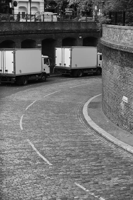 A person wearing a blue jacket and dark trousers is loading large white cardboard boxes into the back of a white removal van parked on a paved city street. The van's rear doors are wide open, revealing an interior lined with wooden panels. Several additional cardboard boxes, some taped and others unwrapped, are placed on the pavement nearby. The street is lined with multi-storey Victorian-style buildings with white facades and ornate balconies, and leafless trees are visible along the sidewalk. The scene is illuminated by natural daylight, with a few parked cars visible in the background. This image illustrates a home relocation process, involving furniture and box loading for a professional removals service, such as provided by Removal Van Kentish Town, supporting efficient moving logistics.