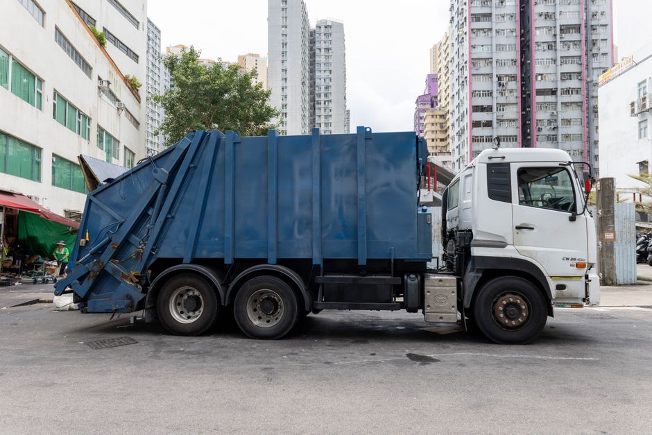 A large green wheeled waste bin with the number 53 and the phrase 'JUN BESA' written on it is positioned on a sidewalk outside a property. The bin is situated next to a shelving unit filled with vario